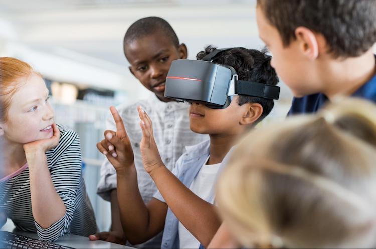 A young student wearing VR goggles and gesturing with his hands, surrounded by 4 other students